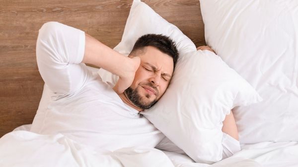 Man suffering with tinnitus in bed covering one ear with his hand and the other with his pillow